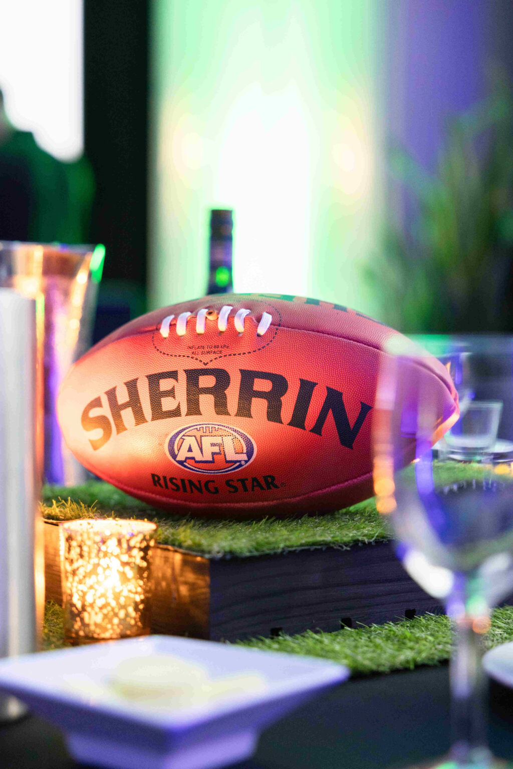 A Sherrin AFL Rising Star football is displayed on a table with decorative grass, surrounded by glassware and a lit candle, with a blurred green and purple background.