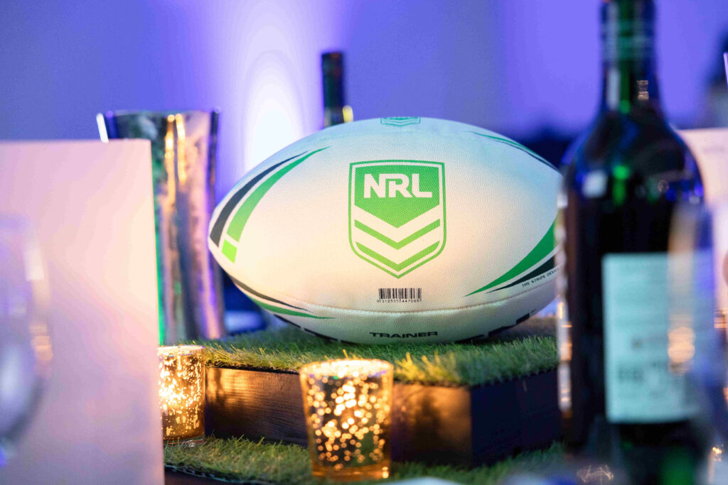 A white and green NRL rugby ball is displayed on a grass-covered centerpiece surrounded by candles and wine bottles, with a blurred blue-lit background.