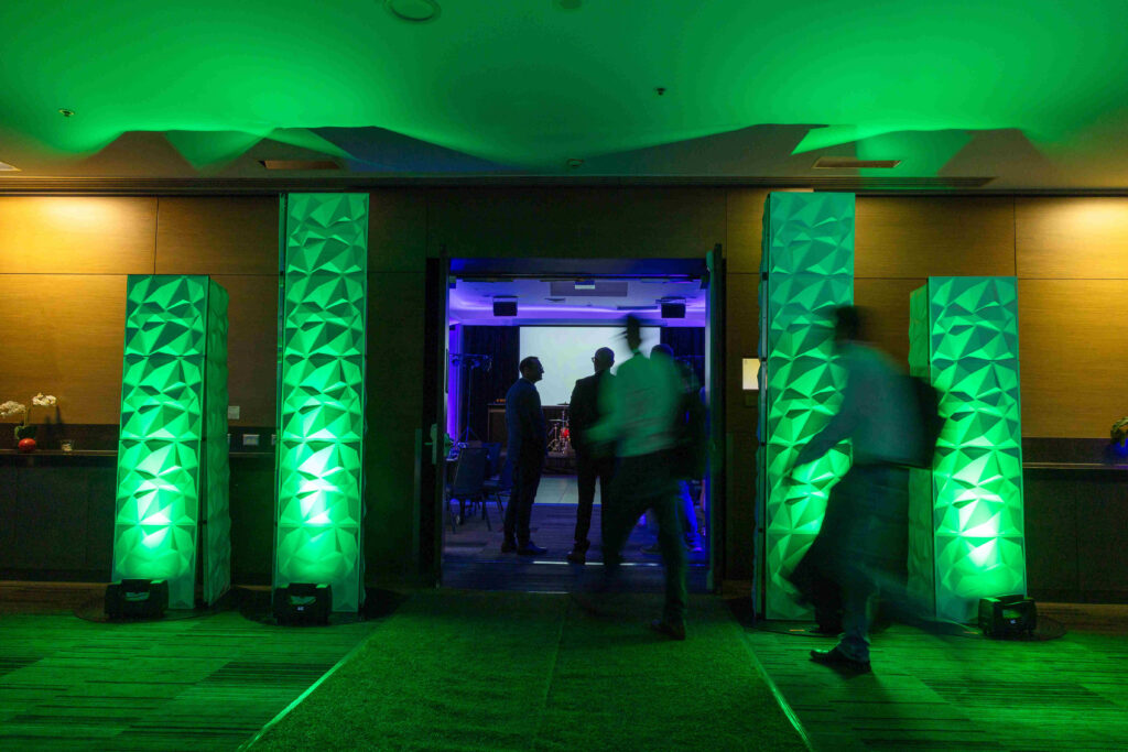 Green-lit decorative columns frame the entrance to an event room, where silhouettes of people are visible inside. Two people walk into the room, creating a dynamic scene with colorful ambient lighting.