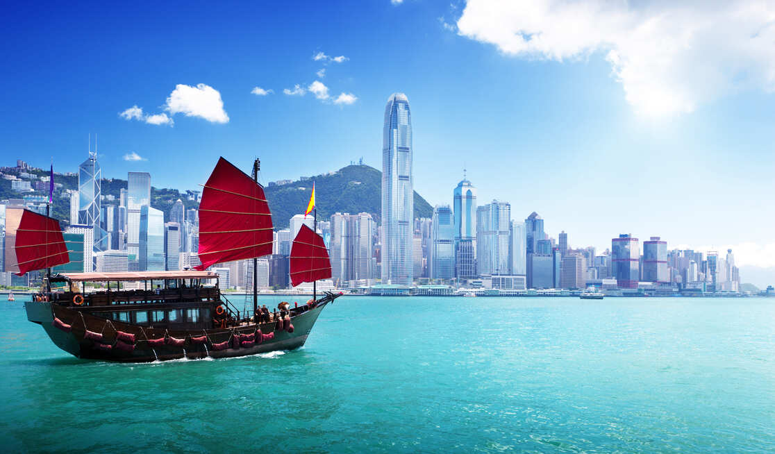 A traditional Chinese junk boat with red sails sails on turquoise water in front of the modern skyline of Hong Kong, with tall skyscrapers and a blue sky with some clouds.