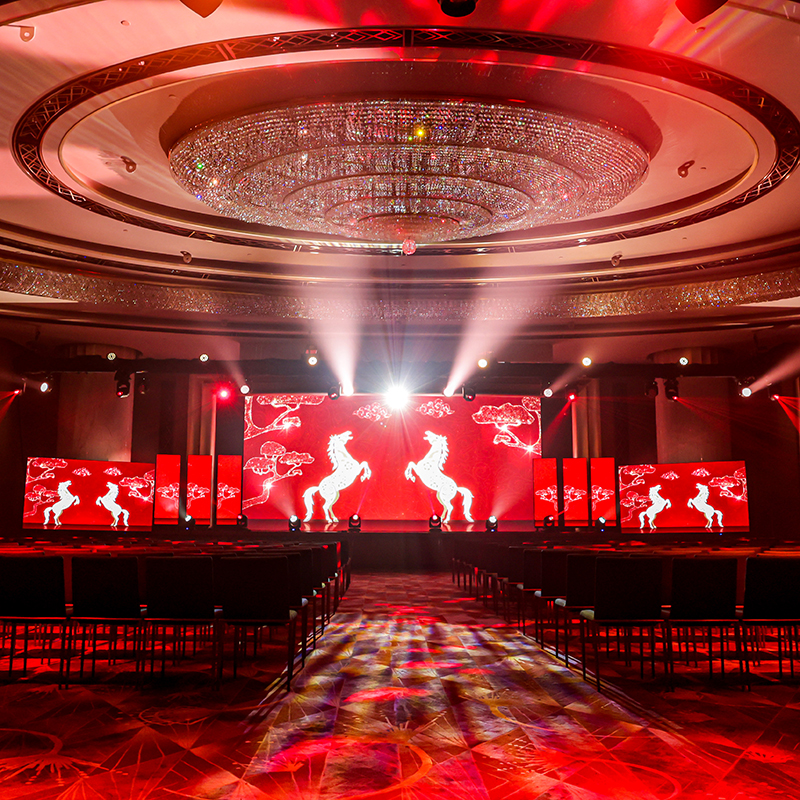 A grand event hall arranged by an event production company in Hong Kong features rows of empty chairs facing a red-lit stage, where large screens display white horse silhouettes and decorative patterns under a sparkling circular chandelier.