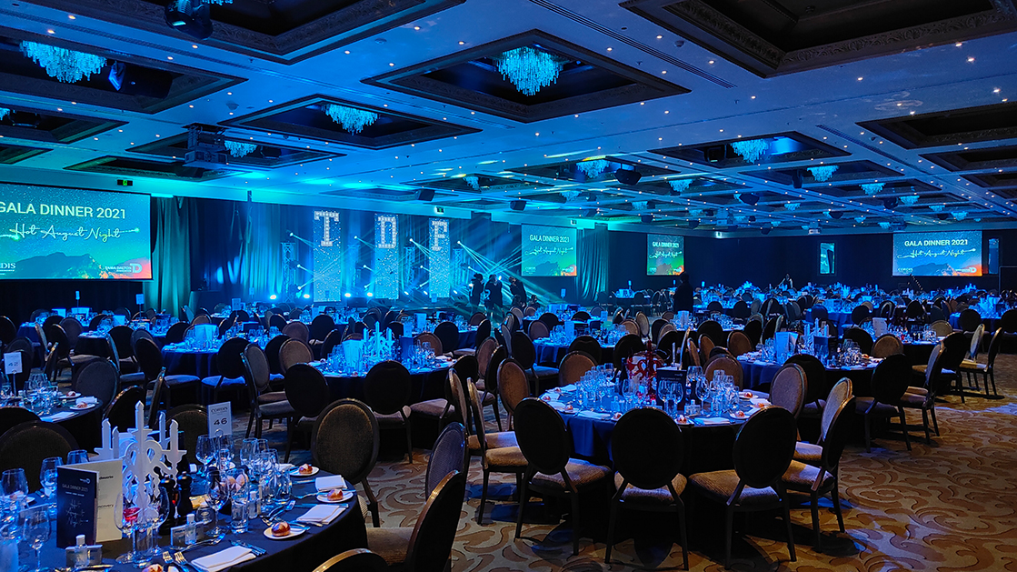 A large, elegant banquet hall is set up for a gala dinner, with blue-lit decorations, glassware, and chairs around round tables. At the front, a stage with blue lighting and event screens showcases expert work by an AV company in Auckland.