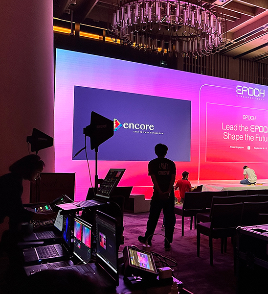 Several people work with audio-visual equipment in a dimly lit room facing a large, colorful display screen that reads Lead the EPOCH, Shape the Future. Chairs are arranged in front of the stage.