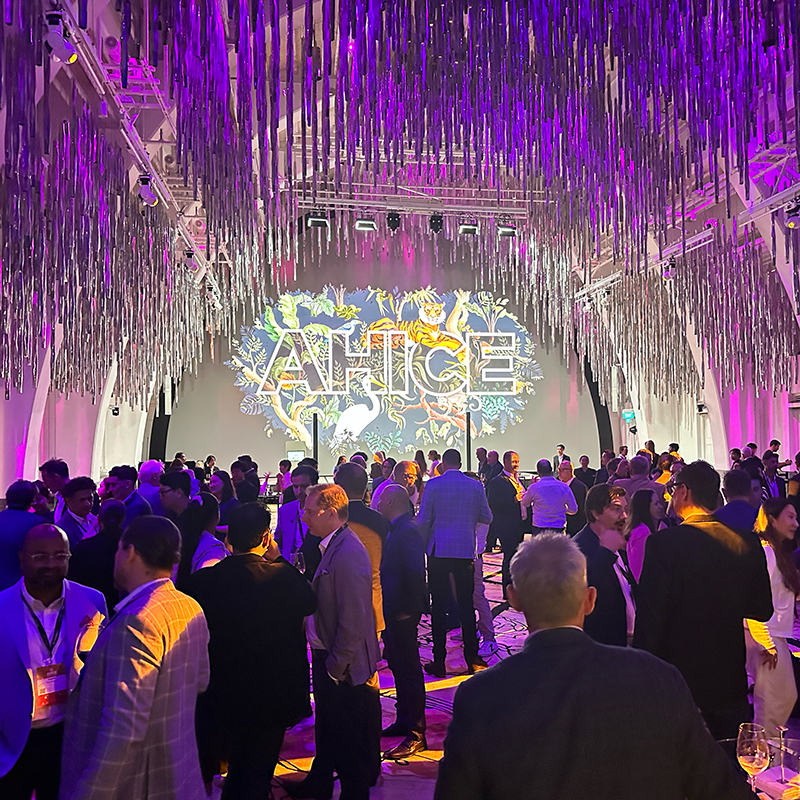 A large crowd of people in formal attire gathers in a vibrant, purple-lit event space with metallic streamers and a colorful AHICE sign projected onstage, showcasing the caliber of event production Singapore is known for.
