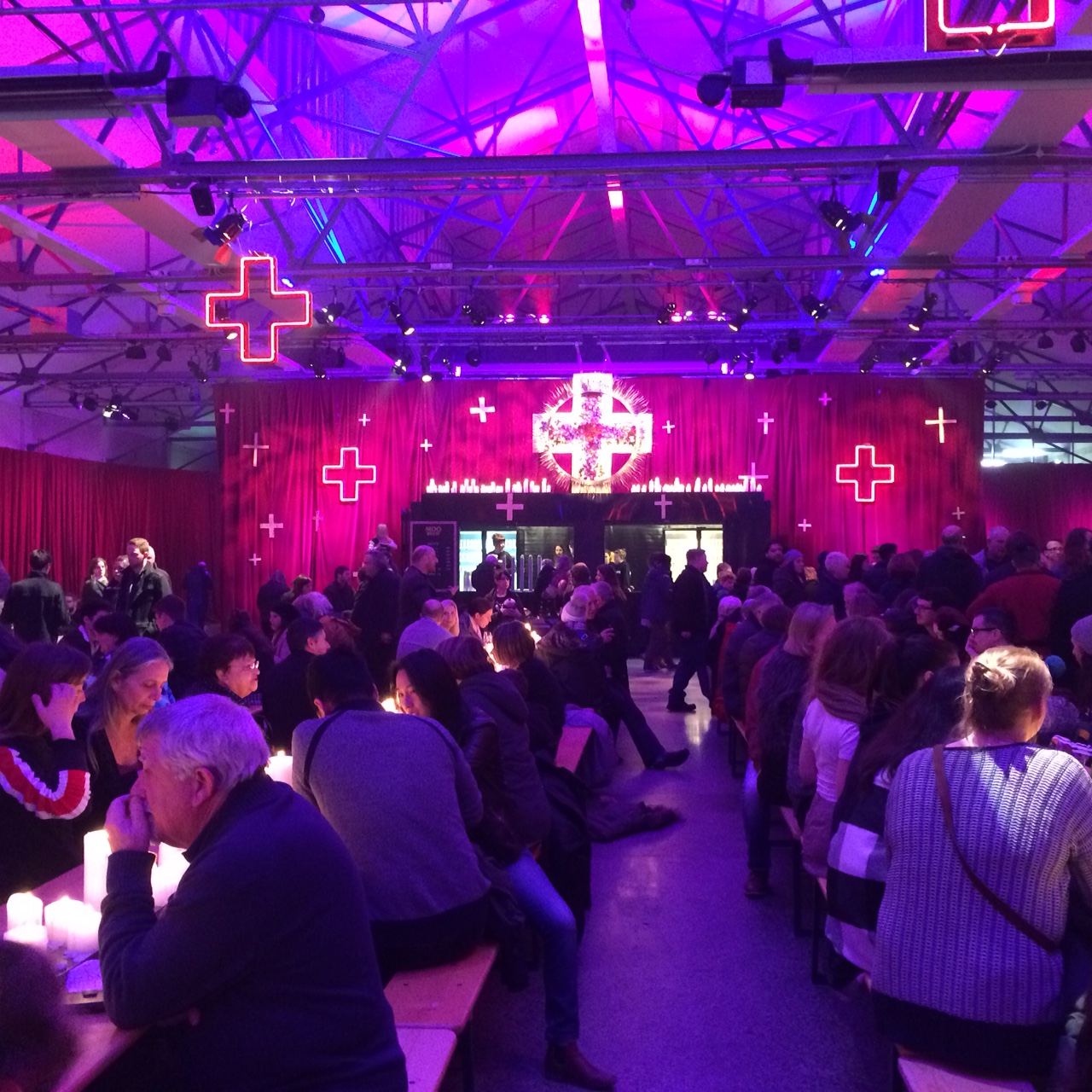 A large crowd gathers in a spacious hall with purple lighting and glowing red cross decorations, expertly designed by event production Hobart specialists. People sit and stand at long tables, socializing in a festive indoor atmosphere.