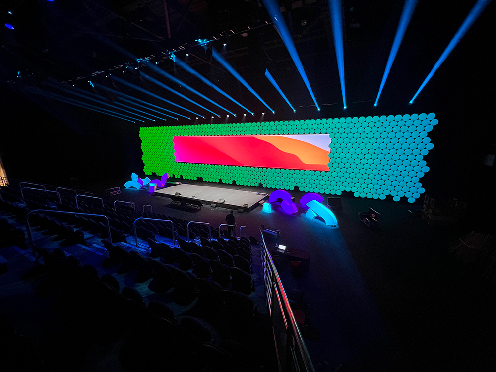 A brightly lit stage with a large, colorful digital screen and a green circular backdrop, surrounded by blue spotlights, in a dark auditorium with empty seats.
