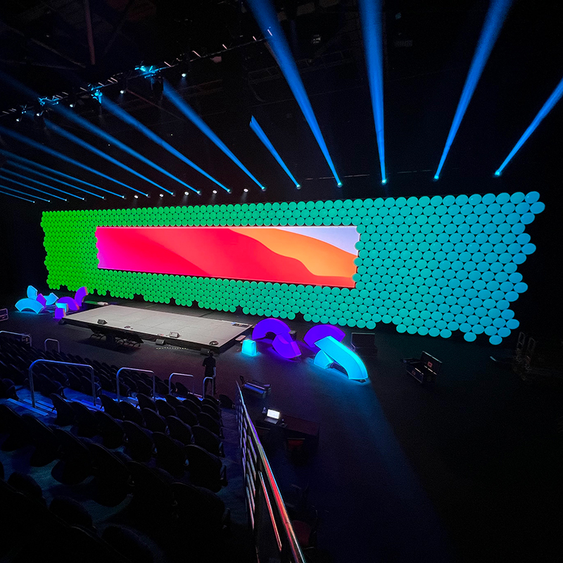 A large, modern stage is illuminated with blue spotlights. The backdrop, crafted by Encore Gold Coast, features a wall of circular green lights and a wide screen displaying a colorful landscape. Rows of empty seats fill the foreground.