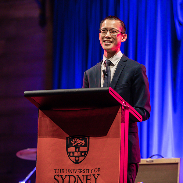 A man in a suit and glasses smiles while speaking at a podium with The University of Sydney logo on it. Blue curtains and stage lights are visible in the background.