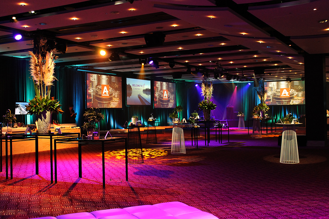 A spacious, modern event hall with tall cocktail tables, floral centerpieces, colorful uplighting, and large projection screens. Elegantly decorated and set up for a formal gathering by a leading AV company Auckland.