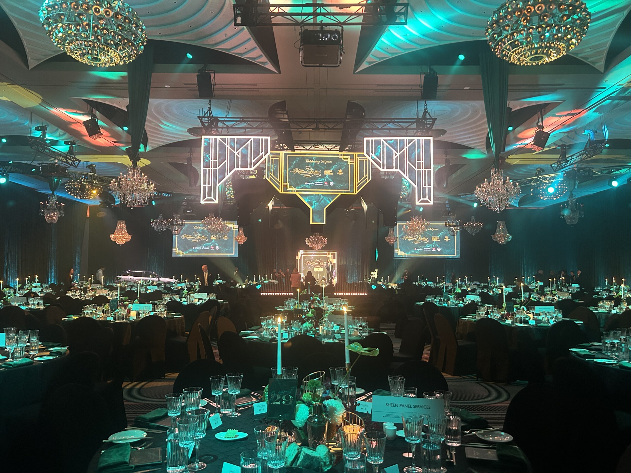 A lavish banquet hall is set for Million Dollar Lunch with round tables elegantly arranged, crystal chandeliers, colorful stage lighting, and glowing neon frames highlighting a decorated stage at the front.