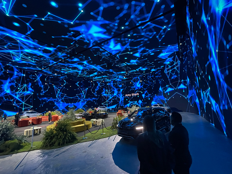 People stand inside a large, immersive room with blue, glowing, digital patterns projected on the walls and ceiling. There are cars, couches, greenery, and the name Amy Shark illuminated in the background.