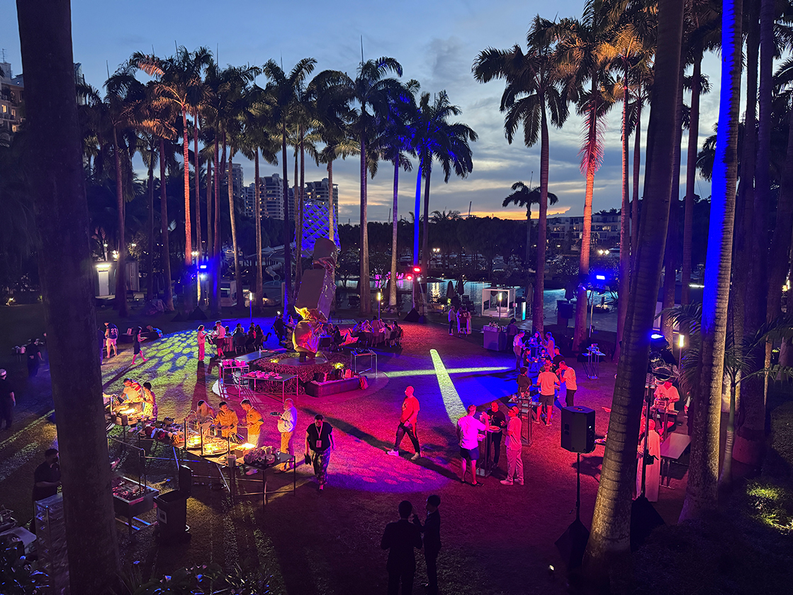 A vibrant outdoor party by an event production company in Singapore takes place at sunset among tall palm trees, with colorful lights illuminating guests mingling, food stations, and tables set on a grassy area. Buildings and a pool are visible in the background.