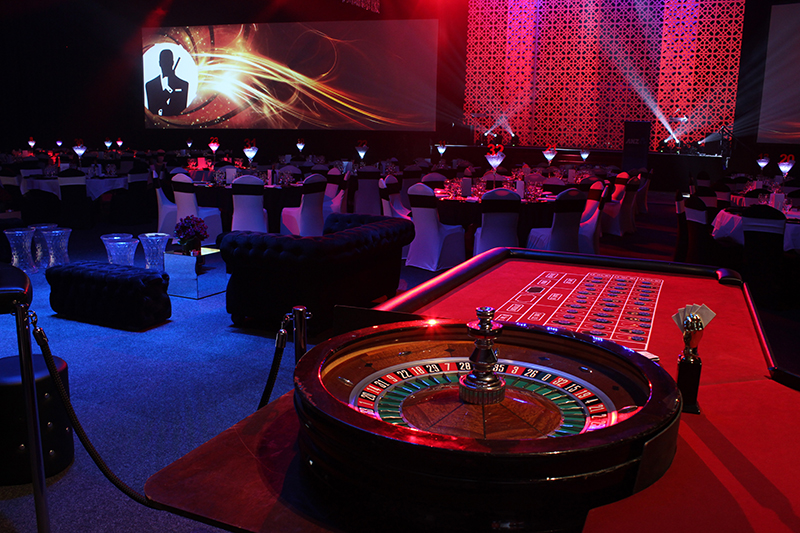 A roulette table in the foreground of a dimly lit, elegant casino-themed event with round tables, black chairs, glowing centerpieces, and a James Bond-themed backdrop.