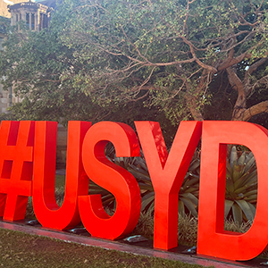 Large red letters forming #USYD are displayed outdoors in front of trees and plants, likely on a university campus.