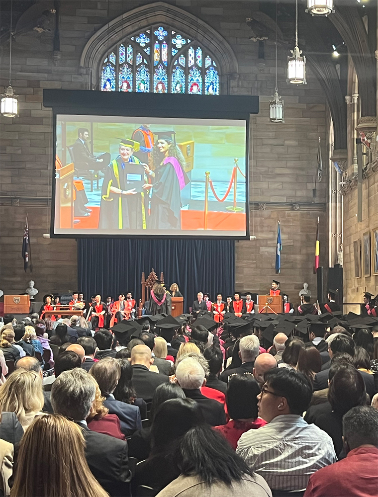 A graduation ceremony in a grand hall with stained-glass windows. Graduates in caps and gowns sit facing a stage where officials are seated. A graduate receives a diploma, shown on a large screen above the stage.