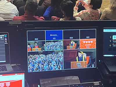 A monitor displays multiple live video feeds of a graduation ceremony, showing speakers at a podium and a crowd of graduates in caps and gowns, while people sit in the audience in the background.