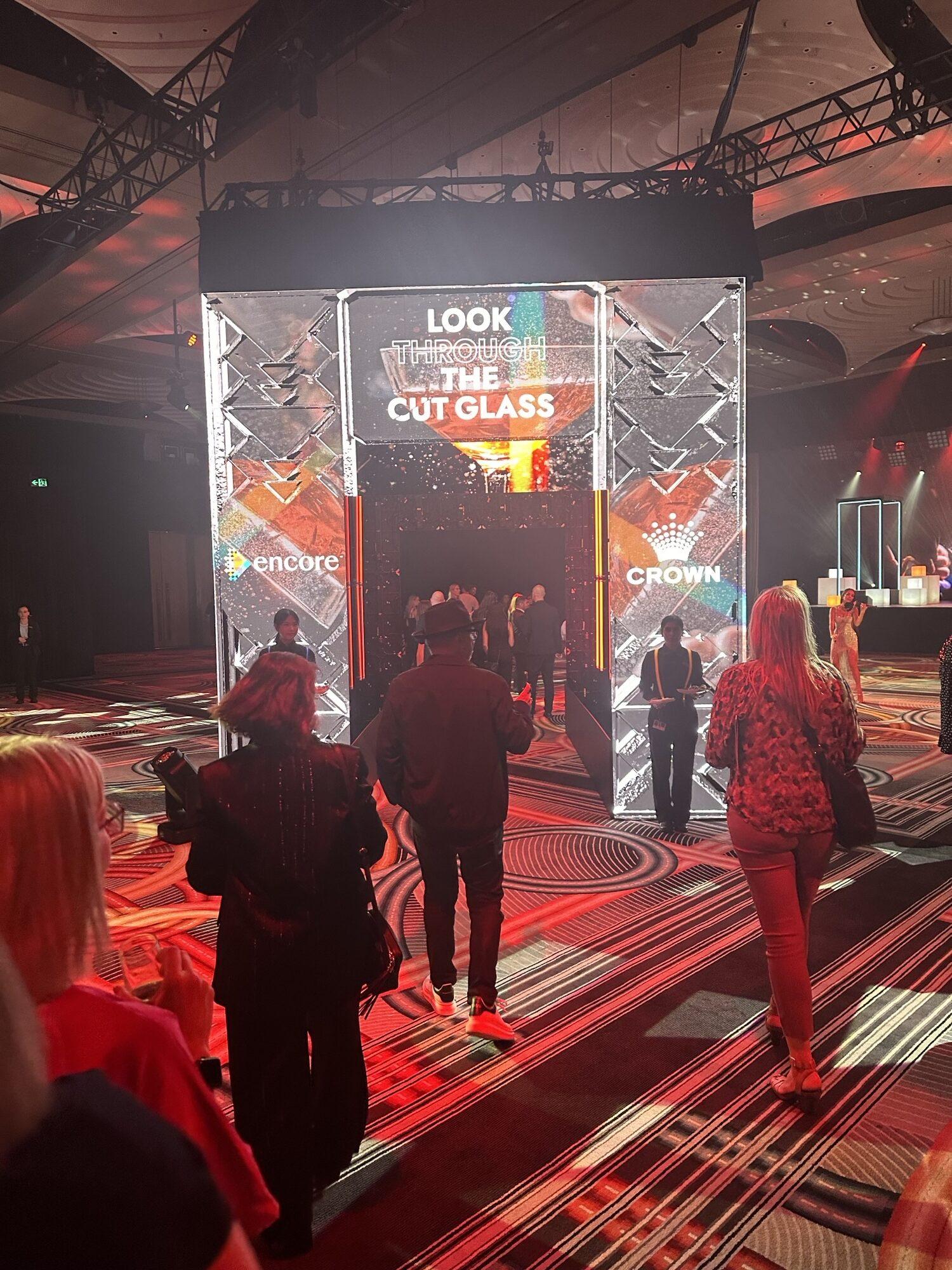 People walk through a brightly lit archway with the words LOOK THROUGH THE CUT GLASS at an indoor event. The venue is decorated with red and black lighting, and logos for Encore and Crown are visible.