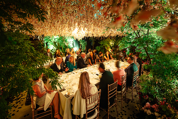 A group of people dressed formally sit around an elegant dinner table under a canopy of hanging flowers and lush greenery, with warm, ambient lighting creating a magical, garden-like atmosphere.