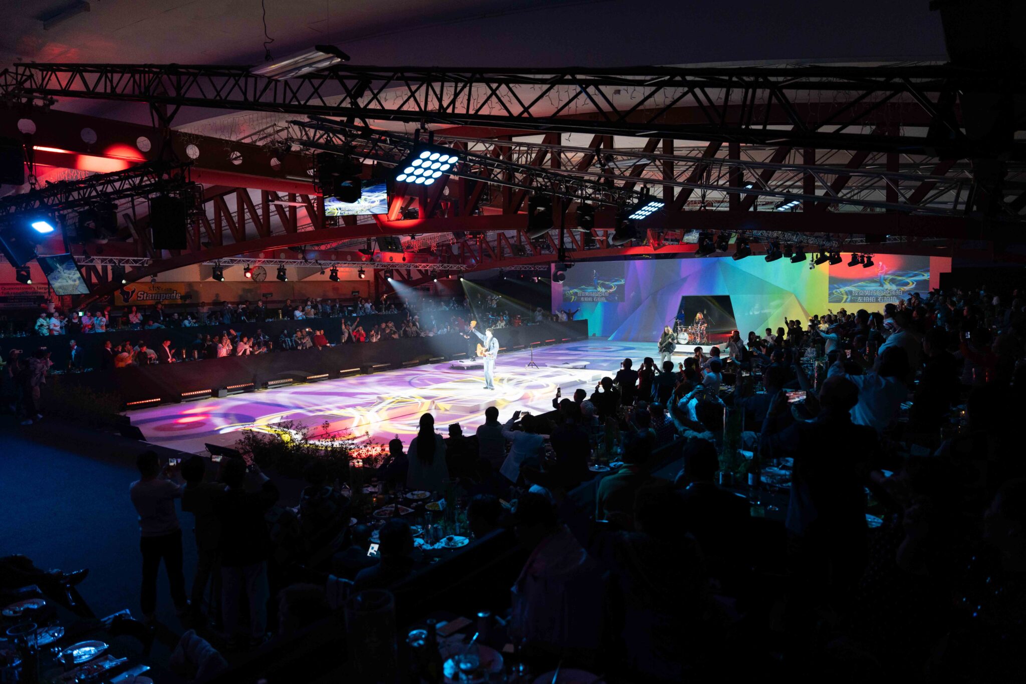A performer stands in the spotlight on a colorful stage inside a large arena, surrounded by an audience seated at tables. The venue is lit with vibrant, multicolored lights creating dynamic patterns on the floor.