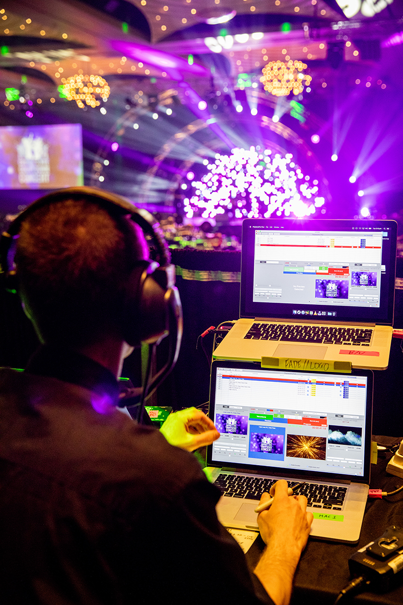 A person with headphones operates two laptops at a control station in a dark, colorful event venue with bright stage lights and digital screens in the background.