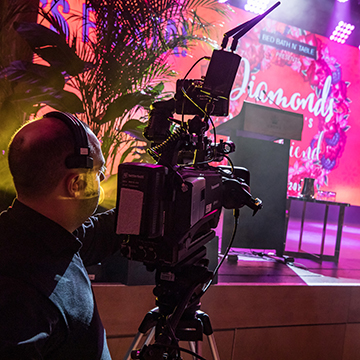 A camera operator with a headset films a brightly lit stage decorated with plants and a colorful sign that reads Diamonds. Event technology and lighting effects combine to create a vibrant atmosphere.