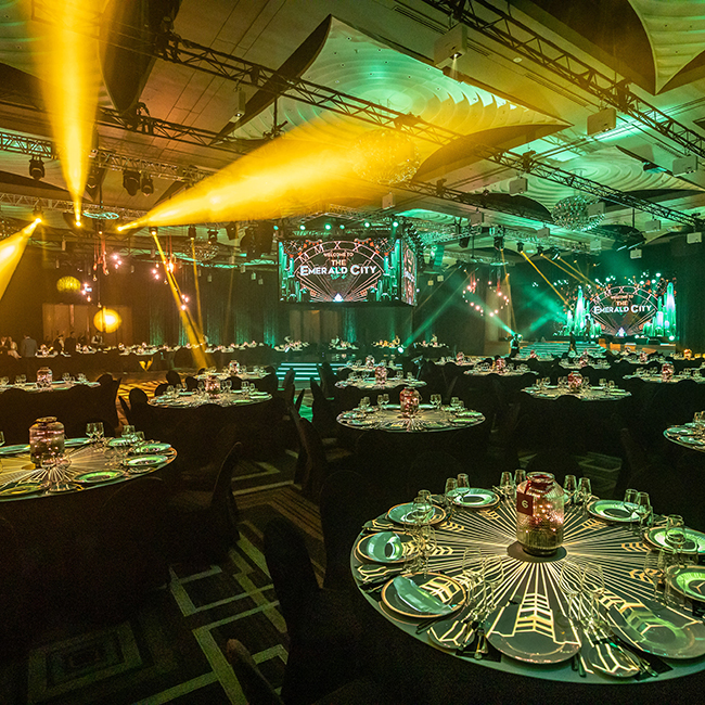A banquet hall set for an event with round tables, elegant place settings, and glowing centerpieces. Yellow stage lights and a large screen reading “Emerald City” create a vibrant, festive Encore atmosphere.