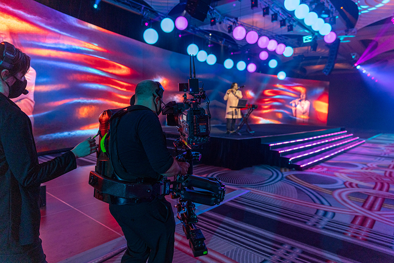 A camera operator films a performer singing on a stage with colorful lights and a large digital backdrop, while another crew member stands nearby. The venue features vibrant patterns and illuminated steps.