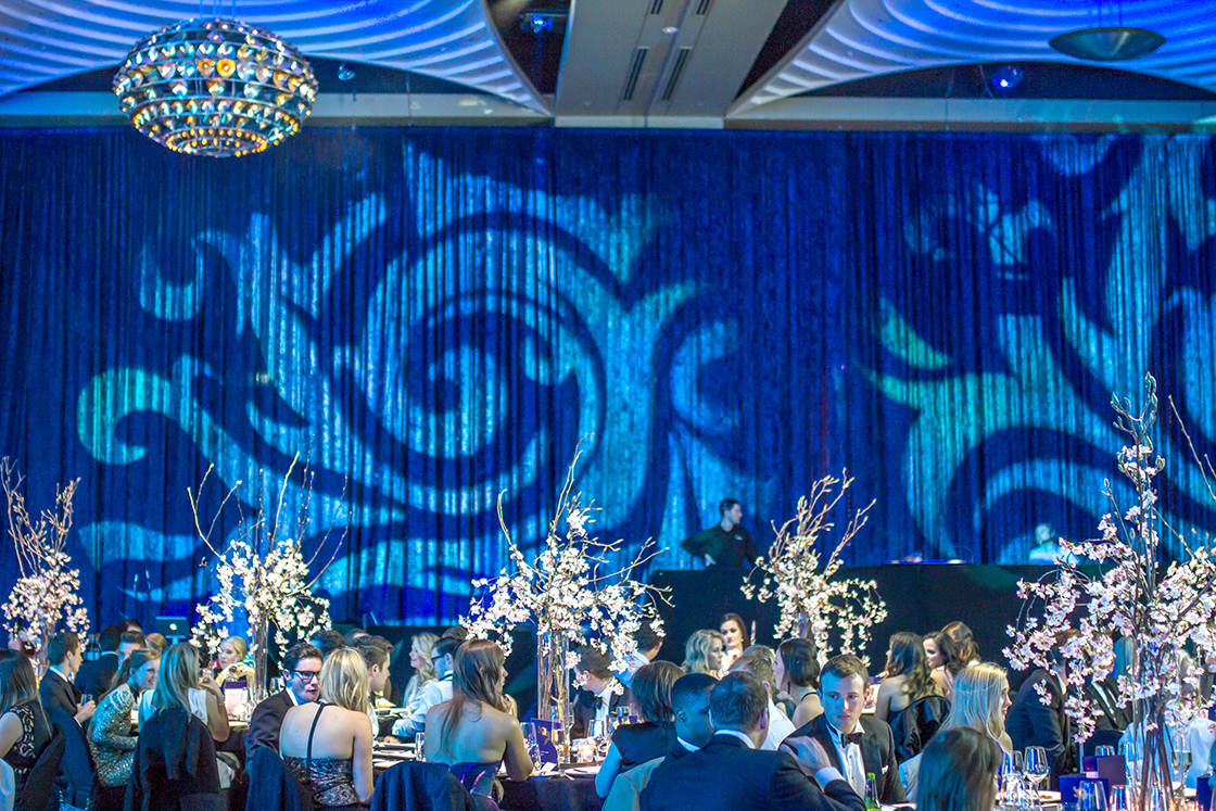 A formal event with guests seated at tables decorated with tall floral arrangements. Blue lighting and swirling patterns are projected onto the walls, creating an elegant atmosphere. A chandelier hangs from the ceiling.