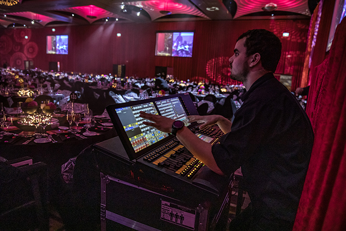 A person operates a lighting control panel at a formal event with round tables, elegant decorations, and dim, colorful lighting. Large screens and seated guests are visible in the background.
