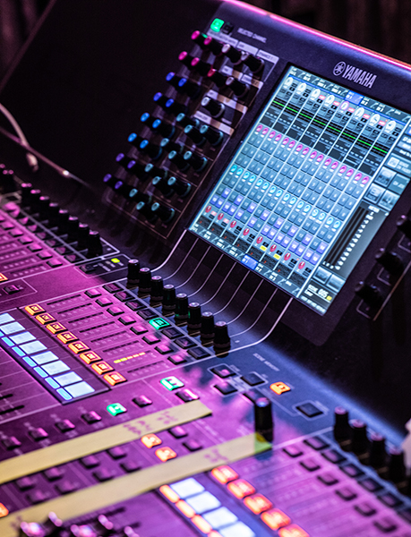 A close-up of a Yamaha digital audio mixing console, showing illuminated buttons, sliders, and a large display screen with various audio channel levels and controls—ready to deliver an unforgettable encore under vibrant stage lighting.