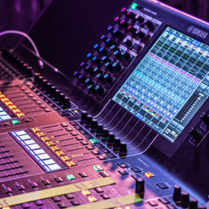 A close-up of a digital audio mixing console, a staple in event technology, with colorful illuminated buttons, faders, and a touchscreen displaying various sound levels and controls.