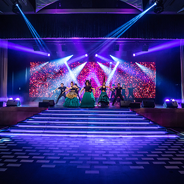 A group of performers in colorful costumes dancing on a stage with vibrant purple and blue lighting, illuminated steps, and a sparkling, colorful background screen.