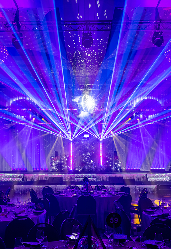A grand event hall is illuminated with vibrant purple and blue stage lights and laser beams, reflecting off a central disco ball. Banquet tables and chairs are arranged in the foreground, ready for guests.