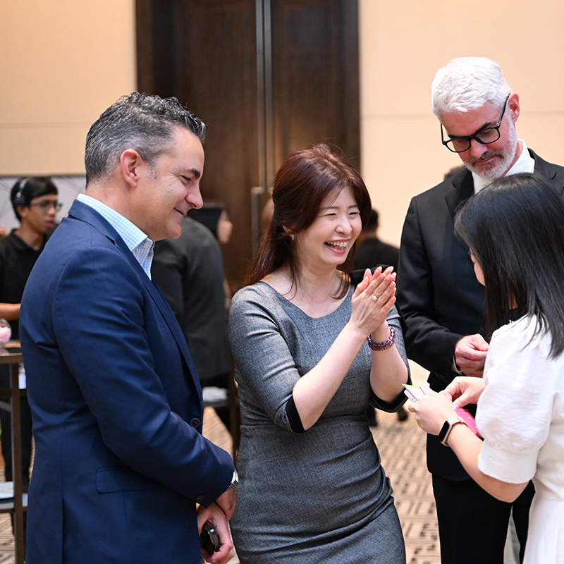 Four adults in business attire stand together indoors, smiling and engaging in conversation. Two women face each other, one holding a name tag, while two men look on, appearing amused and friendly.