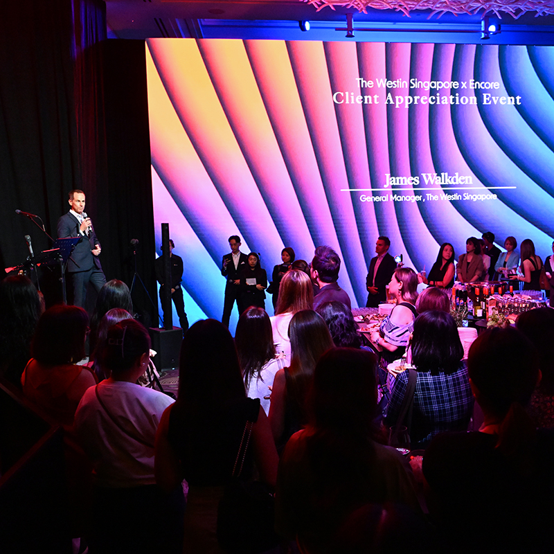 A man in a suit speaks on stage at The Westin Singapore Client Appreciation Event, produced by a leading event production company in Singapore, with a colorful, wave-patterned screen and an audience gathered in a dimly lit room.