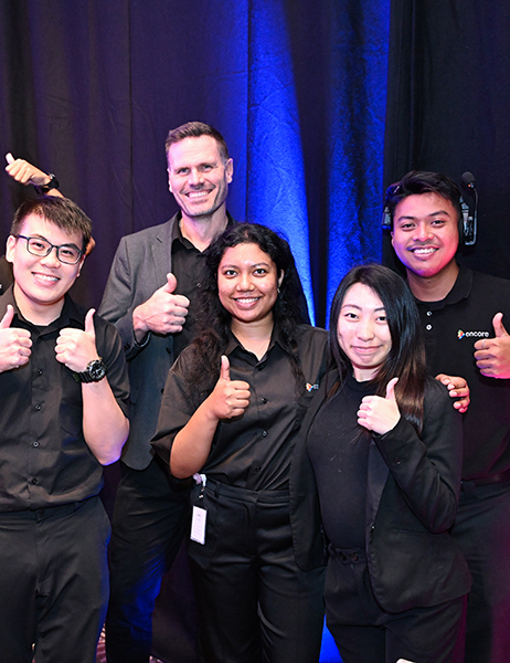 Five people, dressed in black clothing, stand together smiling and giving thumbs up in front of a dark curtain—showcasing the cheerful, enthusiastic team spirit that highlights why work with Encore is such a rewarding experience.