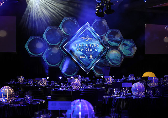 A banquet hall decorated with glowing spherical centerpieces, elegantly set tables, and a stage backdrop featuring hexagonal blue shapes and a sign reading Dine Among the Stars under dramatic lighting.