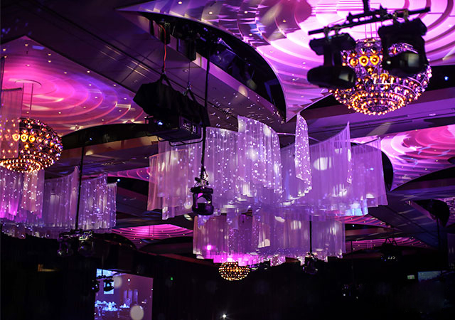 A ceiling decorated with chandeliers and draped, translucent fabric illuminated by purple and pink lights, creating a vibrant, festive atmosphere at an indoor event.