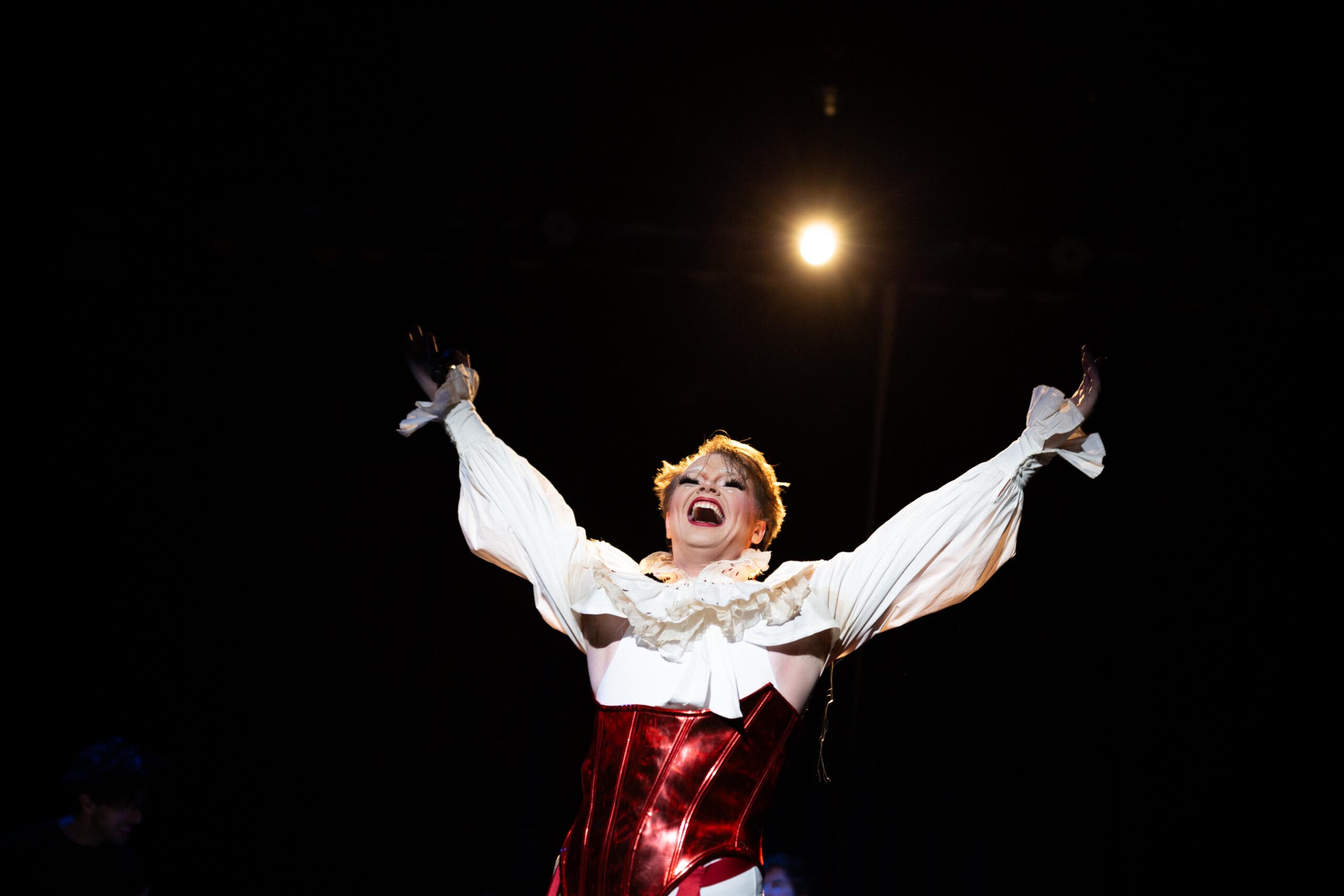 A performer in a dramatic white blouse and shiny red corset stands on stage with arms raised, smiling joyfully under a spotlight against a dark background.