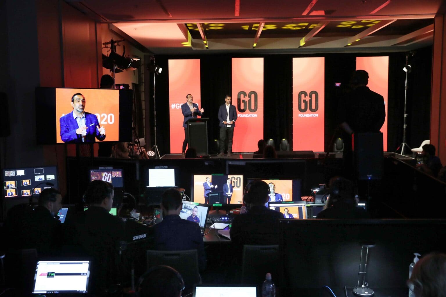A behind-the-scenes view of a live event with people working at computers and control panels, while speakers stand on stage in front of large screens displaying GO FOUNDATION and the number 20.