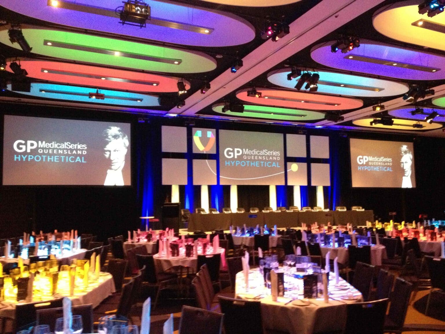 A large event venue with round tables set for dinner, colorful lighting on the ceiling, and multiple large screens displaying GP Medical Series Queensland Hypothetical on stage.