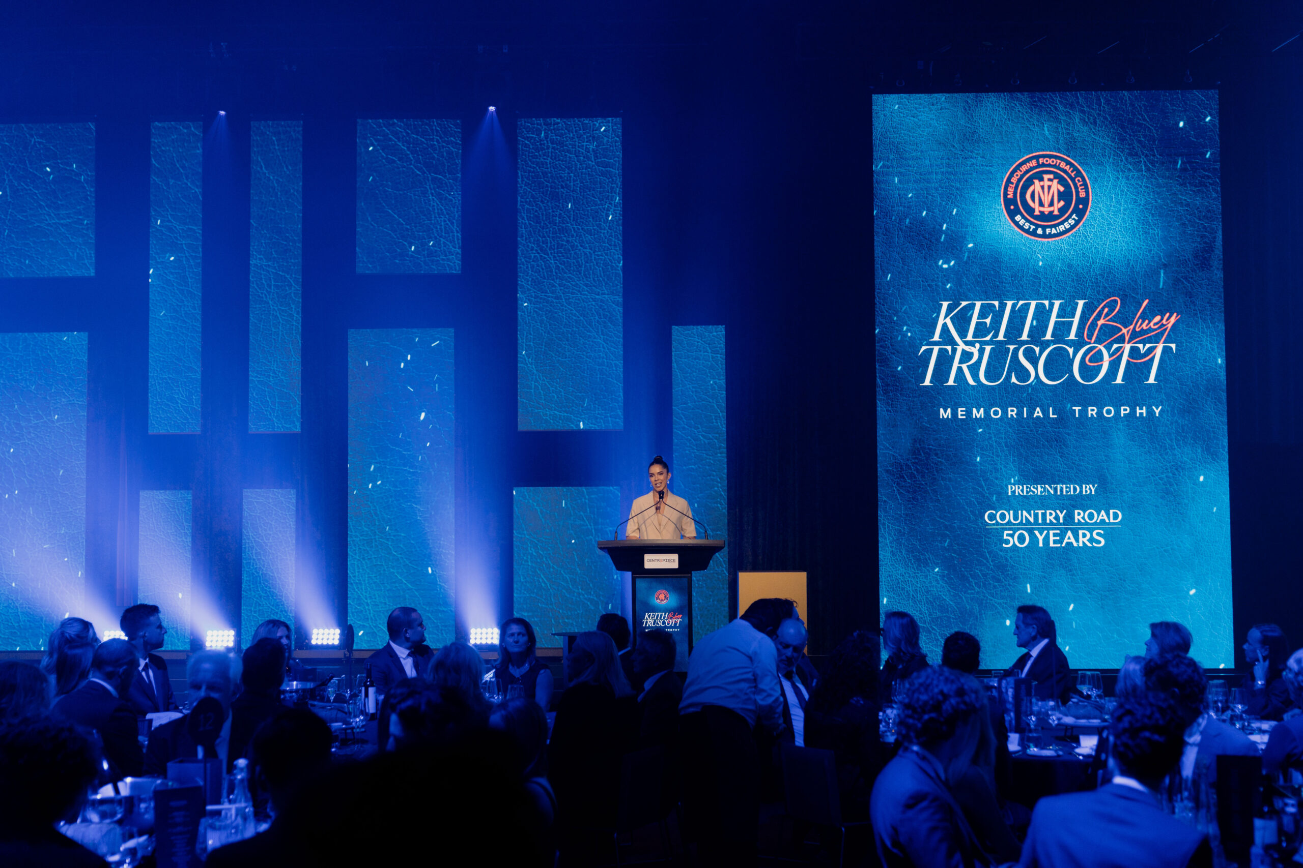 A person stands at a podium on stage under blue lighting at the Keith ‘Bluey’ Truscott Memorial Trophy event, with an audience seated at round tables and a large illuminated event logo displayed behind them.