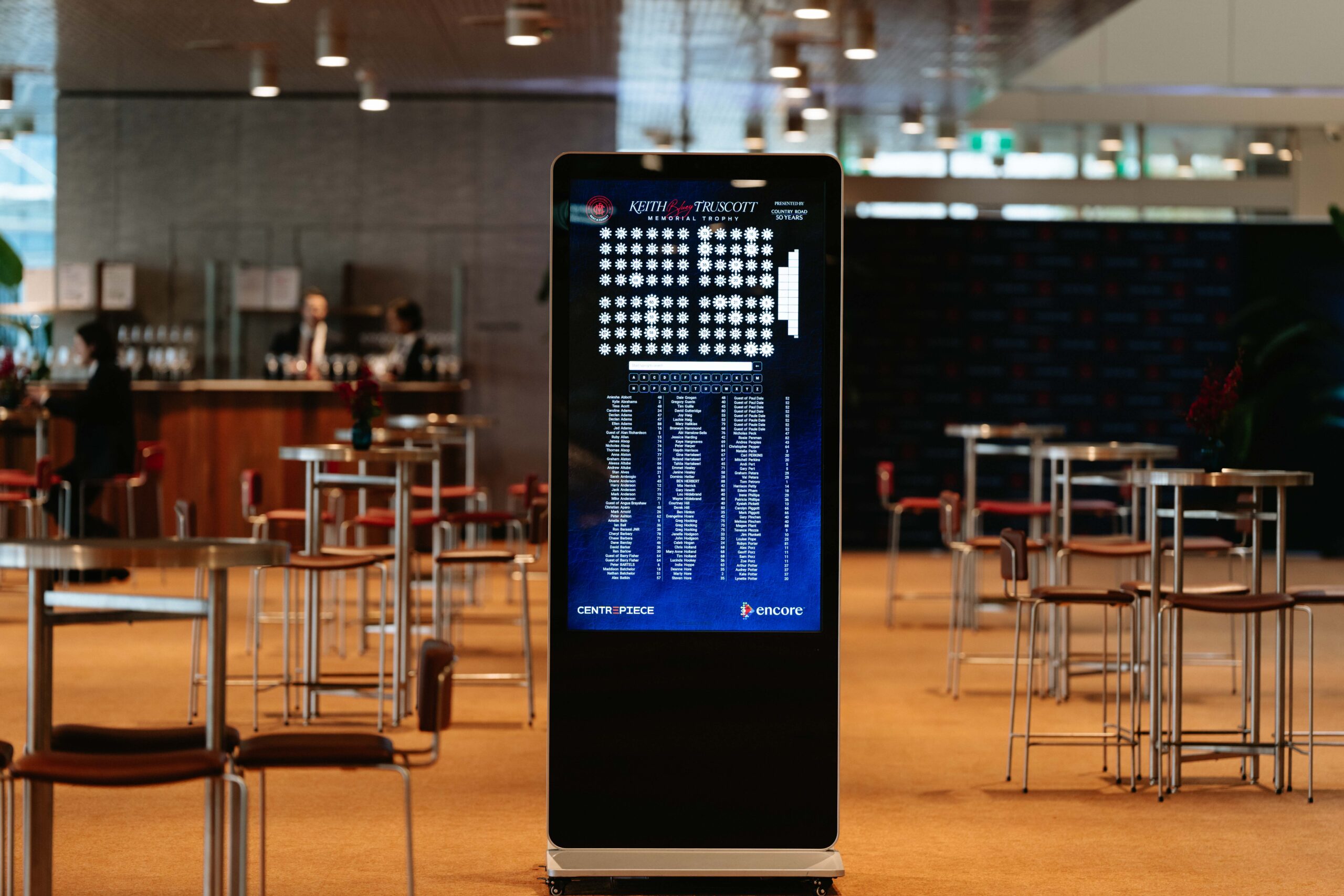 A digital seating chart display stands in the middle of a spacious, modern lobby with empty tables and chairs around it. The screen shows a seating map and a list of names. The background is softly blurred.