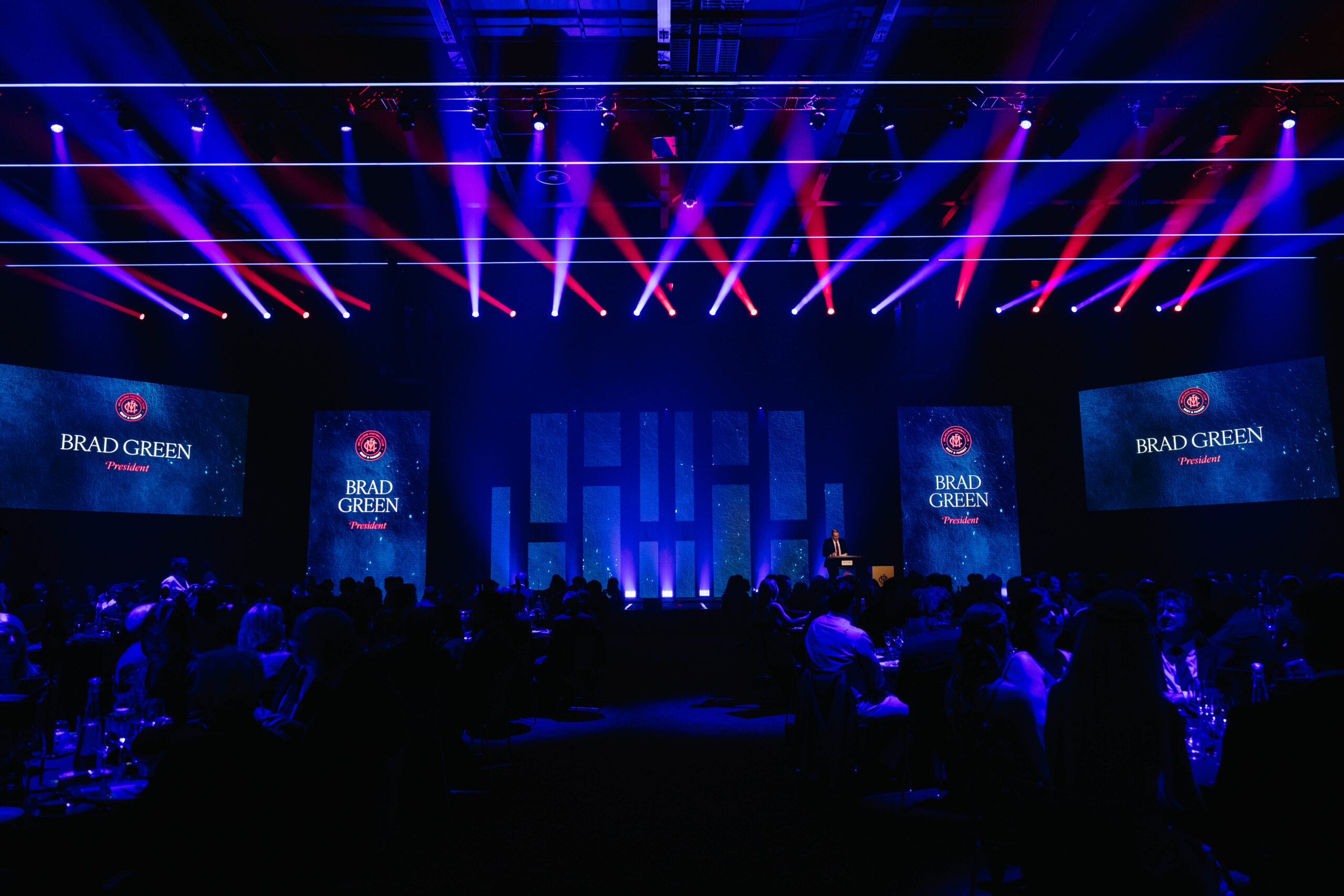 A large, dark event hall filled with seated guests, features colorful stage lighting, multiple screens displaying Brad Green, President, and a central stage with a speaker at the podium.