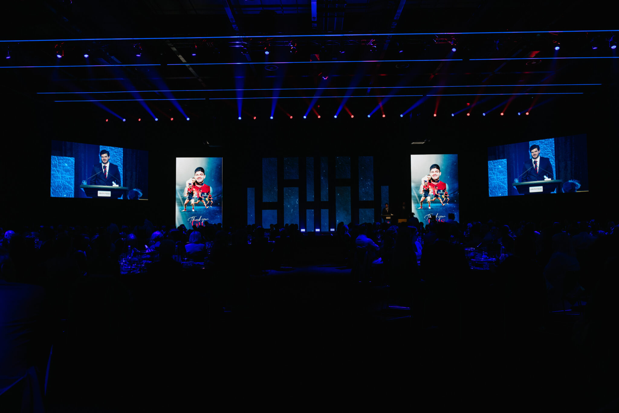 A dark auditorium with an audience faces a stage lit by blue and purple lights. Three large screens display a speaker at a podium and an image of a person in a red outfit.
