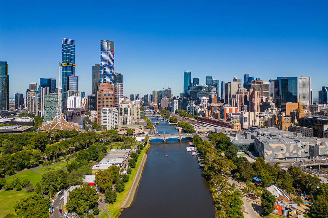 Aerial view of Melbourne’s city skyline with modern skyscrapers, a river running through the center, green parks on both sides, and clear blue skies above.