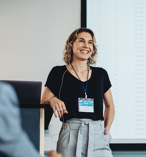A woman in business attire stands at a podium, smiling while giving a presentation about event types. She wears a name badge and headset, with a large screen displaying text or data in the background.