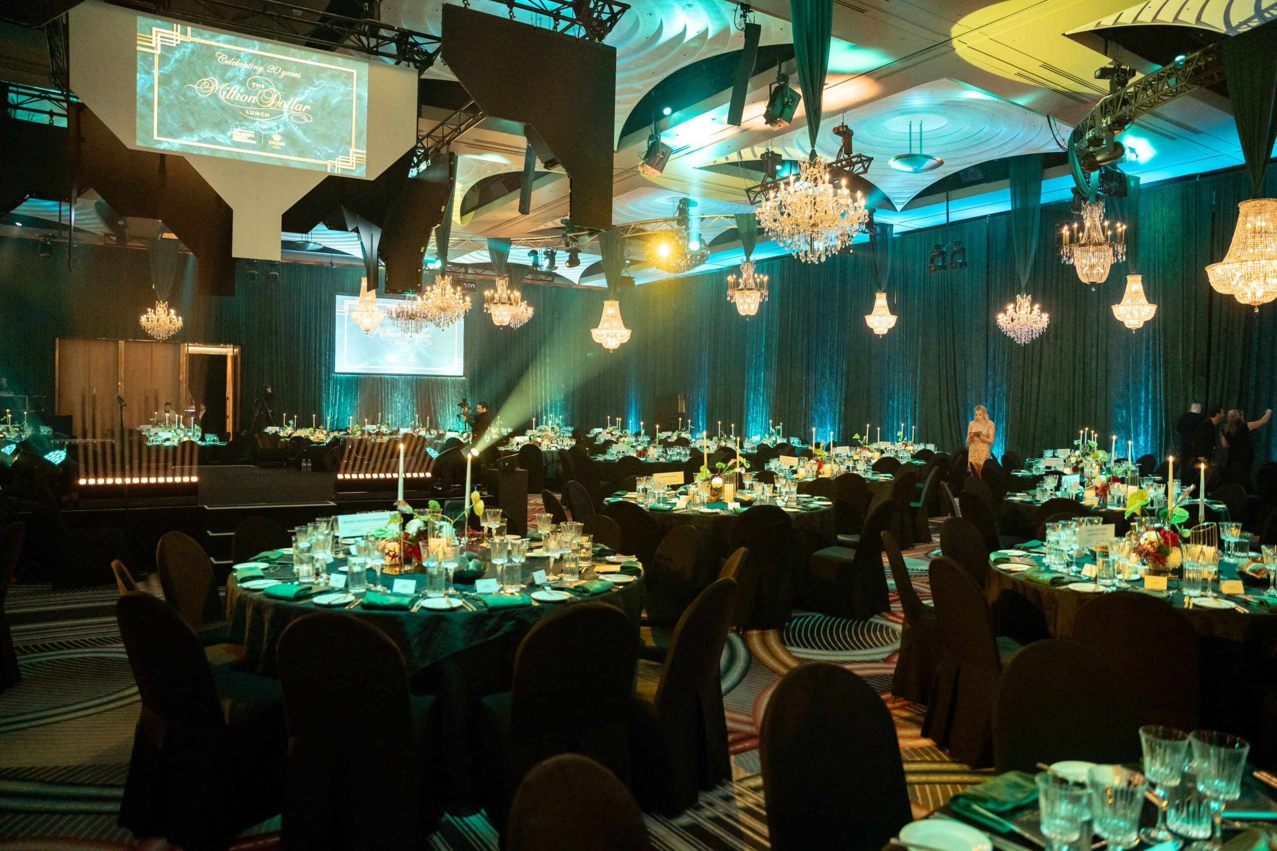 A large, elegant banquet hall set for a formal event at Million Dollar Lunch features round tables, black chairs, chandeliers, and dramatic teal lighting. Tables are set with plates, glasses, and centerpieces, while a stage and screens are visible.