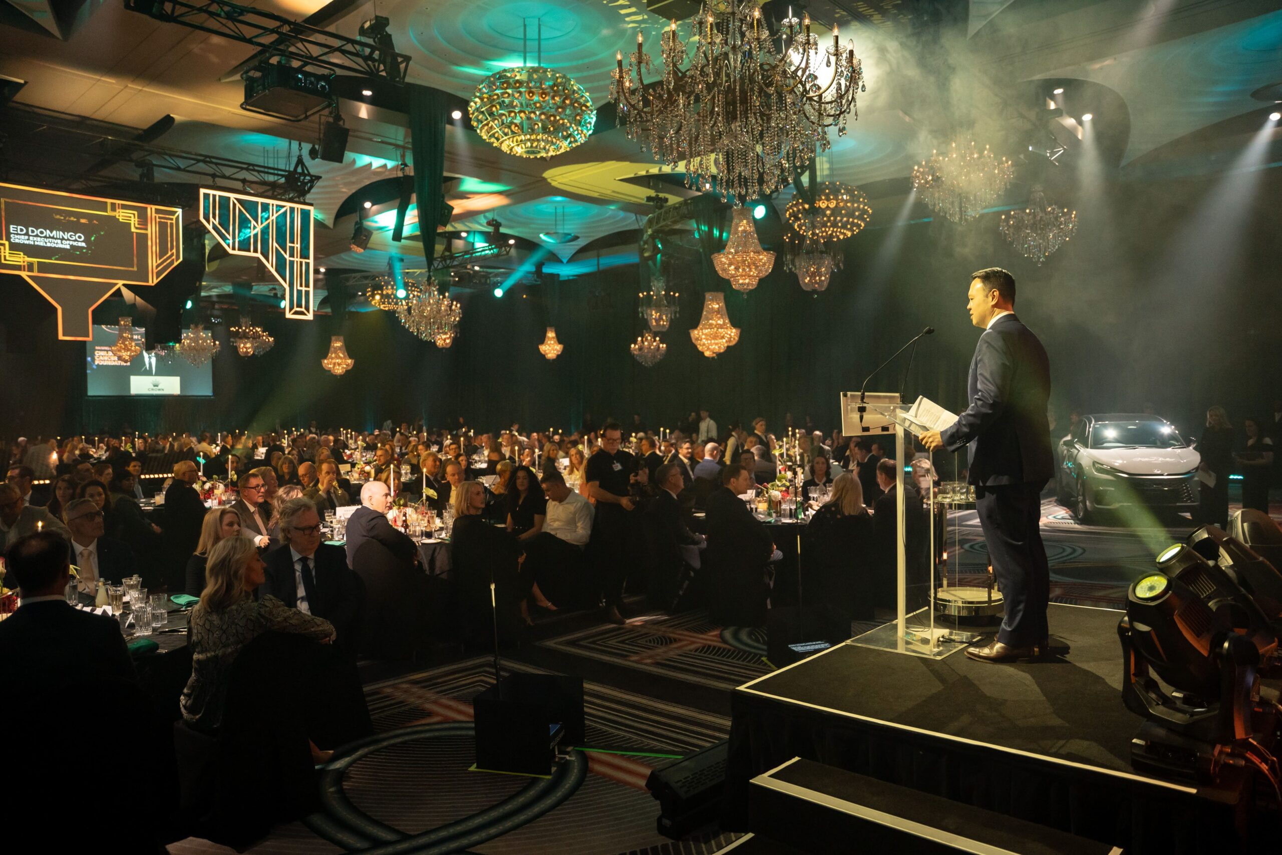 A man in a suit speaks at a podium on a stage at Million Dollar Lunch 2025, addressing a large audience seated at round tables in a grand, chandelier-lit ballroom with green and yellow lighting effects.