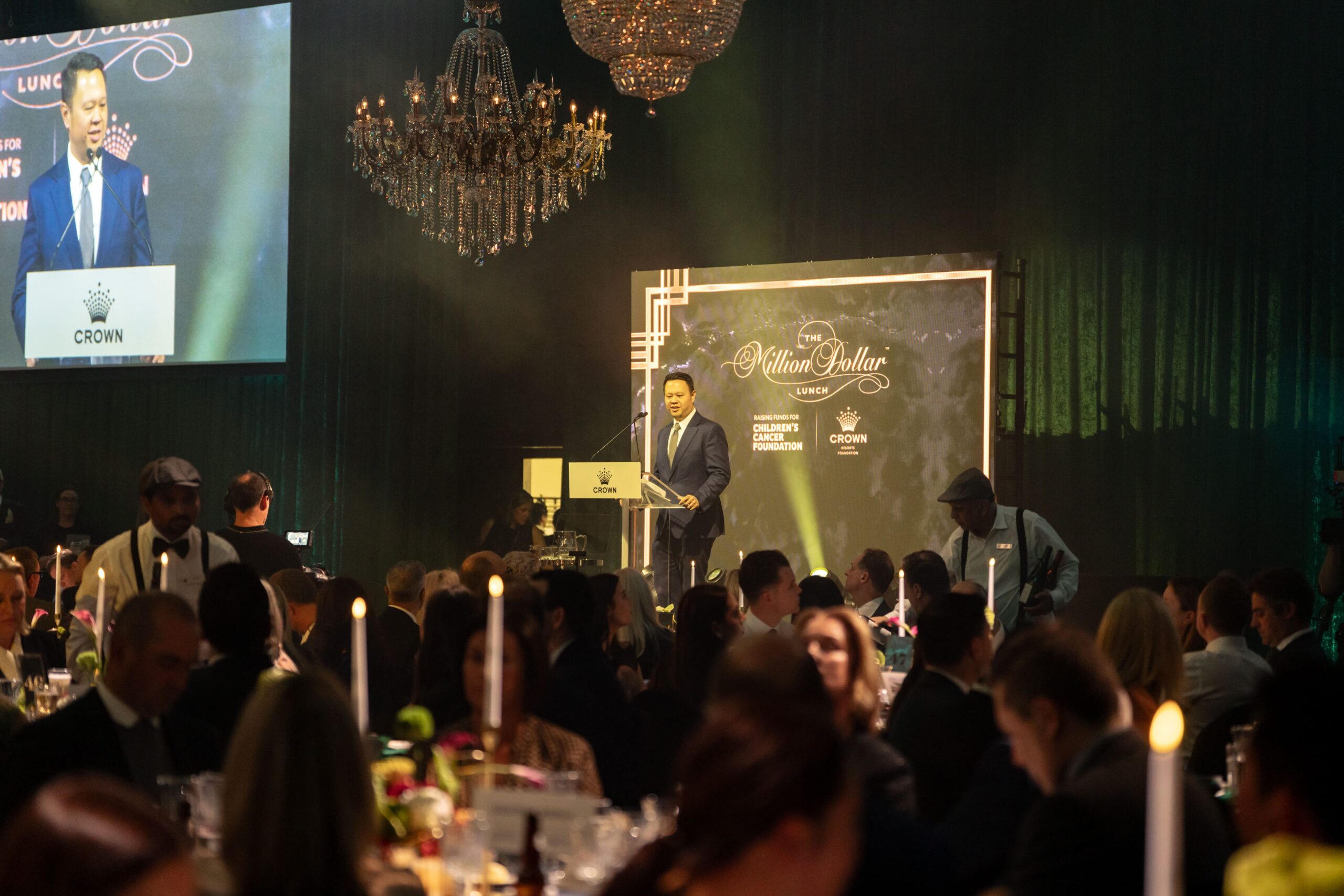 A man in a suit speaks at a clear podium on stage during the formal Million Dollar Lunch 2025 banquet, with a large chandelier overhead and guests seated at candlelit tables in the dimly lit room.
